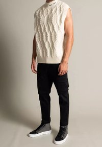 Pletený bezrukávový svetr v krémové barvě s texturovaným diamantovým vzorem, kombinovaný s černými cargo kalhotami a šedými vyššími teniskami.