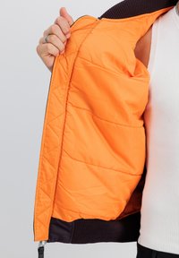 Quiltet jakke med en orange inderside og mørk lilla yderside. Har ribbede mansjetter og lynlåslukning. Glat tekstur, varm polstring.