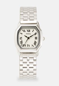 Montre octogonale en argent avec un cadran crème à motifs, des chiffres romains noirs et un bracelet en métal. Le nom de la marque "FOSSIL" est affiché sur le cadran.