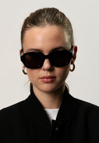 Occhiali da sole neri oversized a forma rettangolare con lenti oscurate, abbinati a un giubbotto bomber nero. Presenta orecchini a cerchio dorati e una camicia bianca sotto.