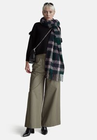 Černý svetr s texturovanými rukávy, zelený a fialový kostkovaný šál s třásněmi, oversized olivové kalhoty s širokými nohavicemi a černé boty s špičatou špičkou.