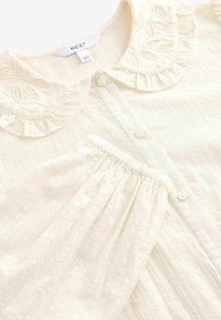 Helle cremefarbene Bluse mit gerüschtem Kragen und bestickten Akzenten, Knopfverschluss und strukturiertem Stoff mit vertikalen Streifen und Punkten.