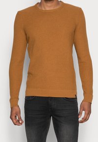 Brun stickad tröja med rund hals, ribbade ärmslut och nederkant. Buren med mörka jeans. Texturen är slät med ett subtilt mönster.