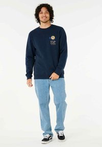 Navy blue sweatshirt with a round neck and long sleeves; features a logo in yellow and white. Paired with light blue jeans and black sneakers.