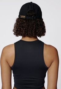 Schwarzes sportliches Crop-Top mit rundem Halsausschnitt und breiten Trägern, kombiniert mit einer schwarzen Kappe, auf der "hummel SPORTSWEAR" in goldener Schrift steht.