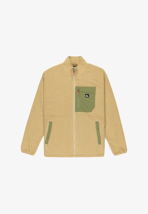 Chaqueta de forro polar beige con cuello alto, cremallera frontal y bolsillo en el pecho verde. Presenta bolsillos laterales y un acabado suave y texturizado.
