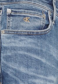 Jasnoniebieskie dżinsy z denimu z złotym przeszyciem, z małym logo „ck” na przedniej kieszeni i metalowymi guzikami.