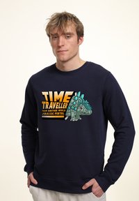 Felpa di colore blu navy con una grafica di un dinosauro stilizzato e il testo "VIAGGIATORE DEL TEMPO" sopra. Tessuto morbido, maniche lunghe.
