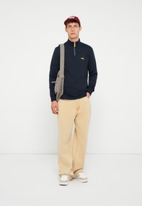 Marine fleece vest met gele hardware, lichtbeige wijde broek, grijze schoudertas en rode pet. Witte sneakers met grijze accenten.
