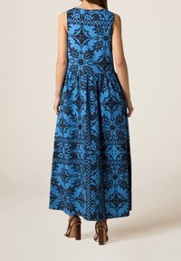 Robe longue bleue avec des motifs floraux et géométriques noirs, sans manches, taille froncée et jupe fluide. Sandales à talons carrés marron.