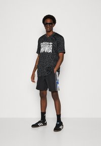 Black graphic t-shirt with white lettering, paired with black shorts featuring side stripes, and black sneakers with white accents. Sunglasses worn.