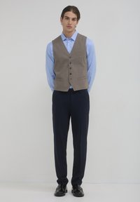 Beige Weste über einem hellblauen Hemd, kombiniert mit dunkelblauen Hosen. Die Weste hat einen Reverskragen und Eingrifftaschen, vervollständigt mit schwarzen Schuhen.