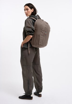 Donna con abbigliamento casual scuro che indossa un grande zaino marrone, in piedi di lato con le mani in tasca contro uno sfondo semplice.