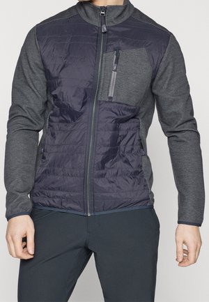 Mężczyzna w granatowej pikowanej kurtce na zamek z szarymi teksturowanymi rękawami i kieszenią na zamek na piersi, w połączeniu z ciemnymi spodniami.