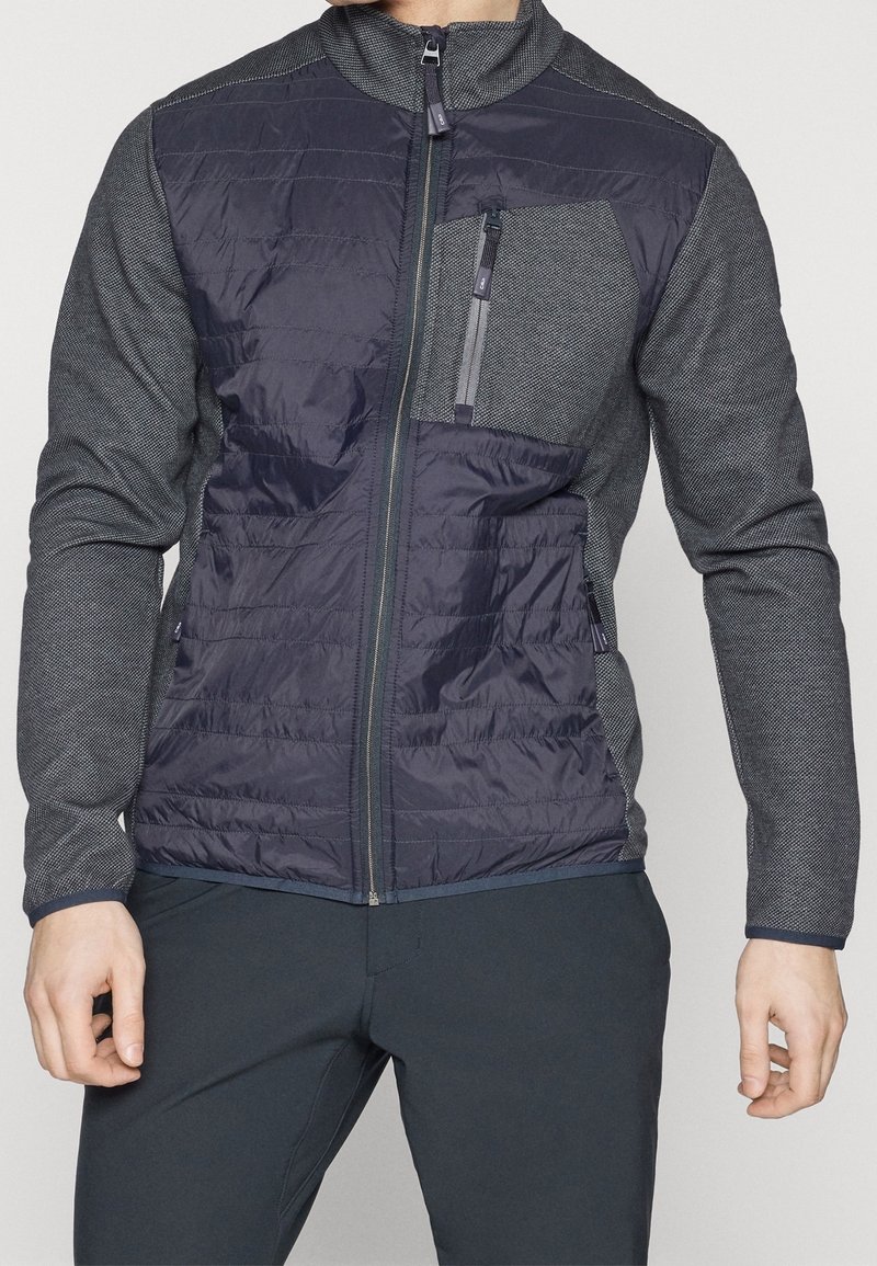 Homme portant une veste matelassée bleu marine zippée avec des manches grises texturées et une poche poitrine zippée, associée à un pantalon sombre.