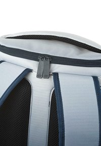 Mochila gris claro con una tela texturizada, que presenta detalles en azul oscuro y un cierre de doble cremallera en la parte superior. Panel de malla visible en las correas.