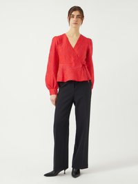 Blouse enveloppante rouge à texture avec de longues manches bouffantes et un détail à nouer, associée à un pantalon noir droit et des escarpins noirs pointus.