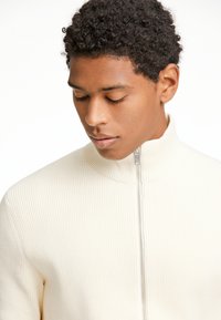 Sudadera cremosa con cremallera, de canalé, con cuello alto y cremallera plateada. Textura suave y silueta ajustada.