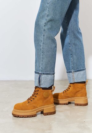 Bottines en daim marron avec une semelle en caoutchouc épaisse, dotées de lacets ronds, d'un onglet au talon et d'un patch logo sur le côté. Portées avec un jean léger bleu retroussé.