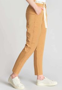 Pantaloni kaki affusolati con cintura regolabile, dotati di tasche laterali, abbinati a sneakers bianche. Tessuto liscio e leggero.