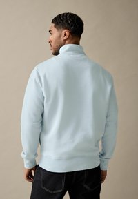 Lichtblauwe sweatshirt met hoge kraag, geribbelde manchetten en tailleband; zachte textuur; gedragen met donkere jeans, weergegeven vanaf de achterkant.