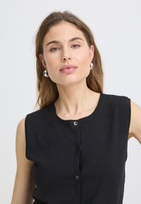 Jeune femme aux longs cheveux châtain clair portant des boucles d’oreilles en argent en forme d’anneaux et un haut noir sans manches boutonné, regardant avec confiance devant elle.