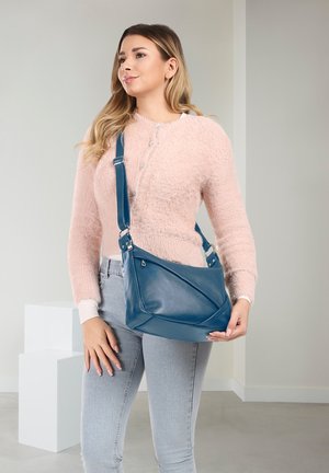 Vrouw draagt een roze pluizige trui en lichte spijkerbroek, houdt een blauwe leren crossbodytas vast in een minimalistische binnenruimte.
