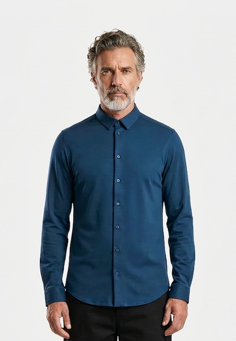 Hallipäine ja habe mees, kes kannab kehast sobivat tumesinist nööbitavat särki ja musti pükse, seisab ühetaolise heleda tausta ees.