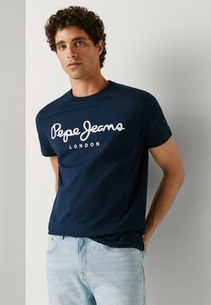 Jaunas vyras su garbanotais plaukais, apsirengęs tamsiai mėlyna „Pepe Jeans London“ marškinėliais ir šviesiai mėlynomis džinsinėmis kelnėmis, stovi priešais paprastą šviesią sieną.