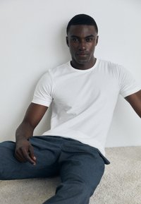 T-shirt blanc à manches courtes en coton, associé à un pantalon à rayures obscures. Modèle assis sur une surface à texture claire.