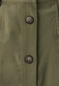 Tissu kaki avec une texture lisse, présentant deux boutons ronds foncés avec un design de visage souriant. Des coutures piquées sont visibles aux côtés des boutons.