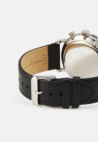 Correa de reloj de cuero negro con costura blanca, hebilla de metal plateado y caja redonda de acero inoxidable visible por el lado. Acabado suave.