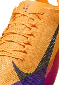 Chaussure de sport orange avec tige en mesh, surface texturée, accents bleus et violets, lacets plats, et logo bien visible sur la languette.