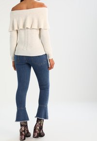 Cremfarbener Off-Shoulder-Pullover mit einem schwingenden Rüschenbesatz, kombiniert mit enganliegenden blauen Jeans und bedruckten Knöchelstiefeln mit einem massiven Absatz.