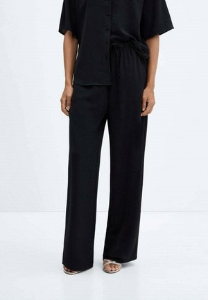 Person trägt schwarze, weit geschnittene Hose und ein lockeres, schwarzes, zum Knöpfen geöffnetes Oberteil, kombiniert mit silbernen Sandalen mit offenen Zehen vor einem weißen Hintergrund.