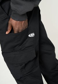 Pantalones cargo negros fabricados con material duradero, con múltiples bolsillos, presentando un destacado parche de logo blanco y detalles de costura en contraste.