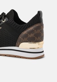 Black sneaker with knit upper, brown textured heel featuring a logo pattern, and gold metallic accent. White midsole with black stripe detail.
