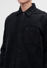 Homme portant une chemise boutonnée en velours côtelé noir avec une poche poitrine et un col pointu, visible des épaules jusqu'au milieu du torse.