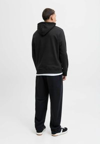 Sort hættetrøje med en glat tekstur og en afslappet pasform, parret med løstsiddende mørke bukser og sorte sneakers med hvide detaljer.