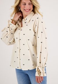 Crèmekleurig katoenen blouse met zwarte strikpatronen, knoopsluiting aan de voorkant en lange mouwen met gathered manchetten en strikdetails.