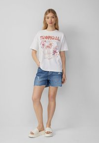 T-shirt graphique blanche avec impression "Tropical", shorts en denim avec bords effilochés et sandales à plateformes beiges. Tenue d'été décontractée.