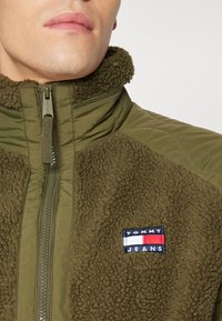 Chaqueta de cremallera verde oliva con textura de forro polar, panel acolchado, cuello alto y un parche con el logo de Tommy Jeans en el lado izquierdo del pecho.