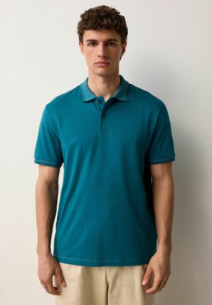 Camisa polo en tono azul verdoso con un cuello estampado. Mangas cortas, textura suave y dos botones en el cuello. Diseño informal combinado con pantalones beige.