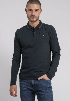 Homme aux cheveux courts portant un polo à manches longues noir avec une bordure jaune et un jean bleu, debout avec une main dans la poche.