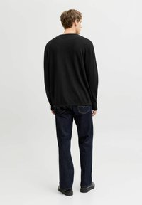 Homme portant une chemise noire à manches longues, un jean bleu foncé et des chaussures noires, debout face à un fond blanc uni.