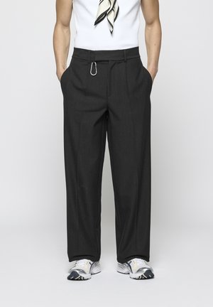 Personne portant un pantalon large noir à fines rayures avec une pince argentée, des baskets blanches et un haut blanc avec une écharpe noire et blanche.