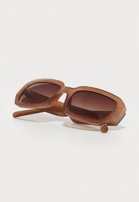 Gafas de sol marrones y de gran tamaño con un marco geométrico, lentes oscuros y una pronunciada línea de las cejas. El material es plástico brillante.