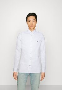 Λευκό πουκάμισο με κουμπιά, φτιαγμένο από βαμβάκι, με κλασικό γιακά, μακριά μανίκια και ένα μικρό λογότυπο στο στήθος. Συνδυάζεται με ανοιχτό μπλε τζιν.