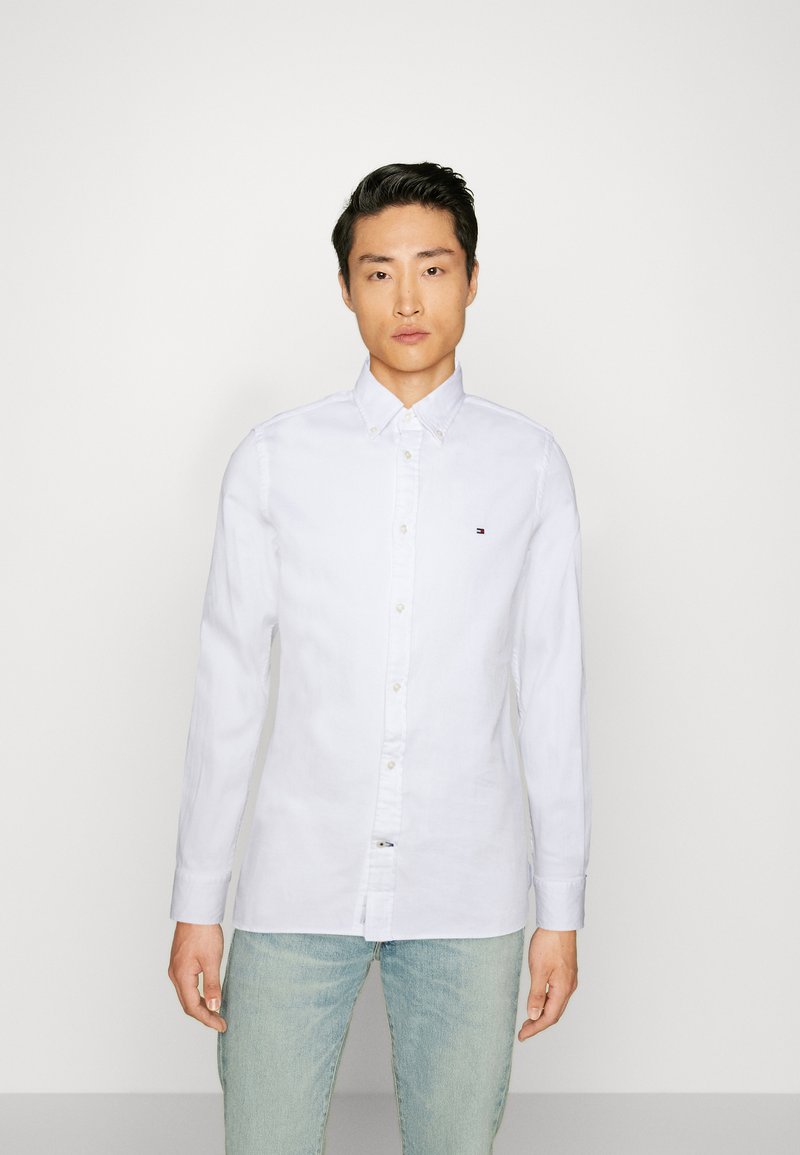 Λευκό πουκάμισο με κουμπιά, φτιαγμένο από βαμβάκι, με κλασικό γιακά, μακριά μανίκια και ένα μικρό λογότυπο στο στήθος. Συνδυάζεται με ανοιχτό μπλε τζιν.