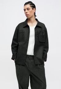 Juodas tekstūruotas marškinėlių švarkas su krūtinės kišene, derinamas su tamsiai žaliais kordūro marškiniais. Modelis vilki baltą maikutę po švarku.